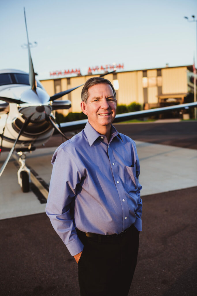 Dwight Holman standing outside in blue collared shirt with plane in the background